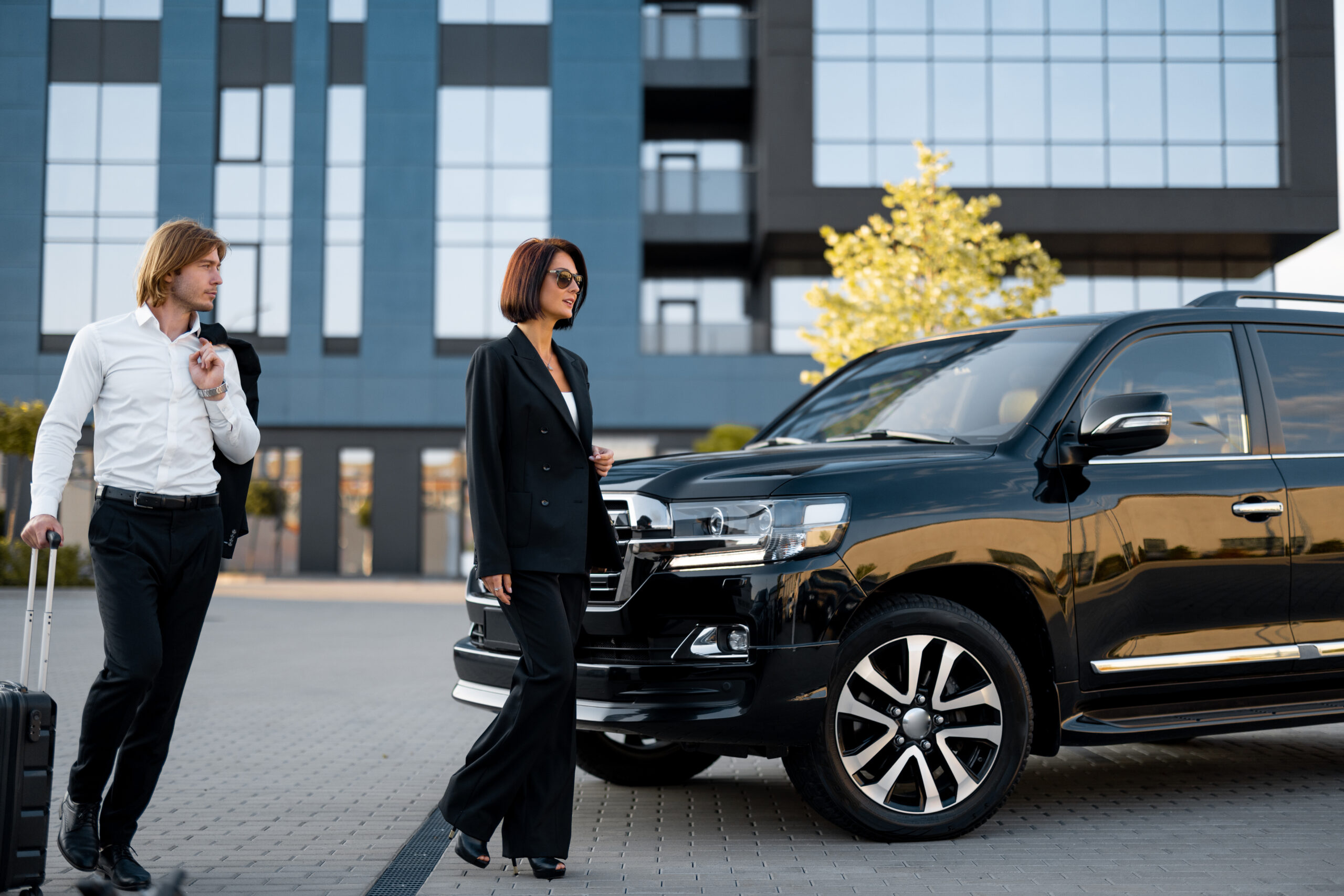 Businessman and businesswoman walk with a suitcase to a luxury black car near hotel or office building, having a business trip together. Concept of transportation and business travel